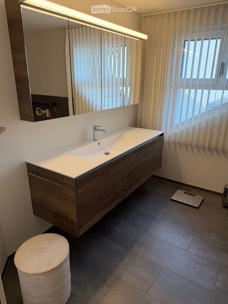 Modern bathroom with a white sink, wooden cabinet, large mirror, wicker stool, and a digital scale on the floor.