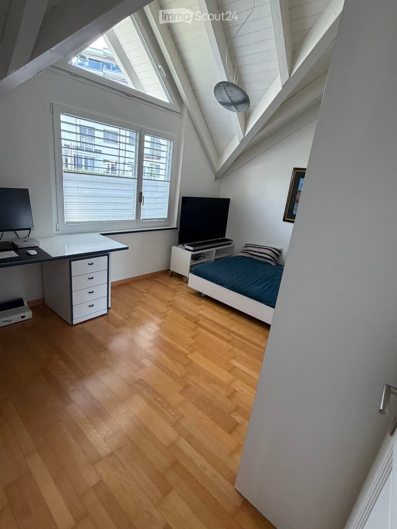 Bright bedroom with wooden flooring, a bed with blue bedding, a desk with a computer, and large windows with blinds.