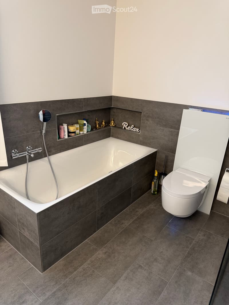 Modern bathroom with a bathtub, toilet, and a shelf holding various toiletries and decorative items. The walls are tiled in dark gray, and the floor is covered with dark gray tiles.