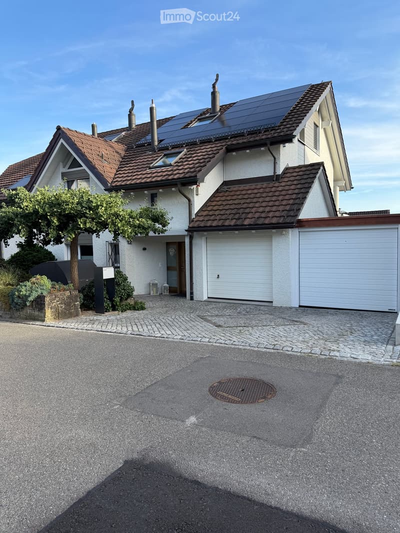 Two-story house with a brown tiled roof, solar panels, white exterior walls, and a garage with a white door. The driveway is paved with cobblestones, and there is a small garden area with a tree and shrubs on the left side.