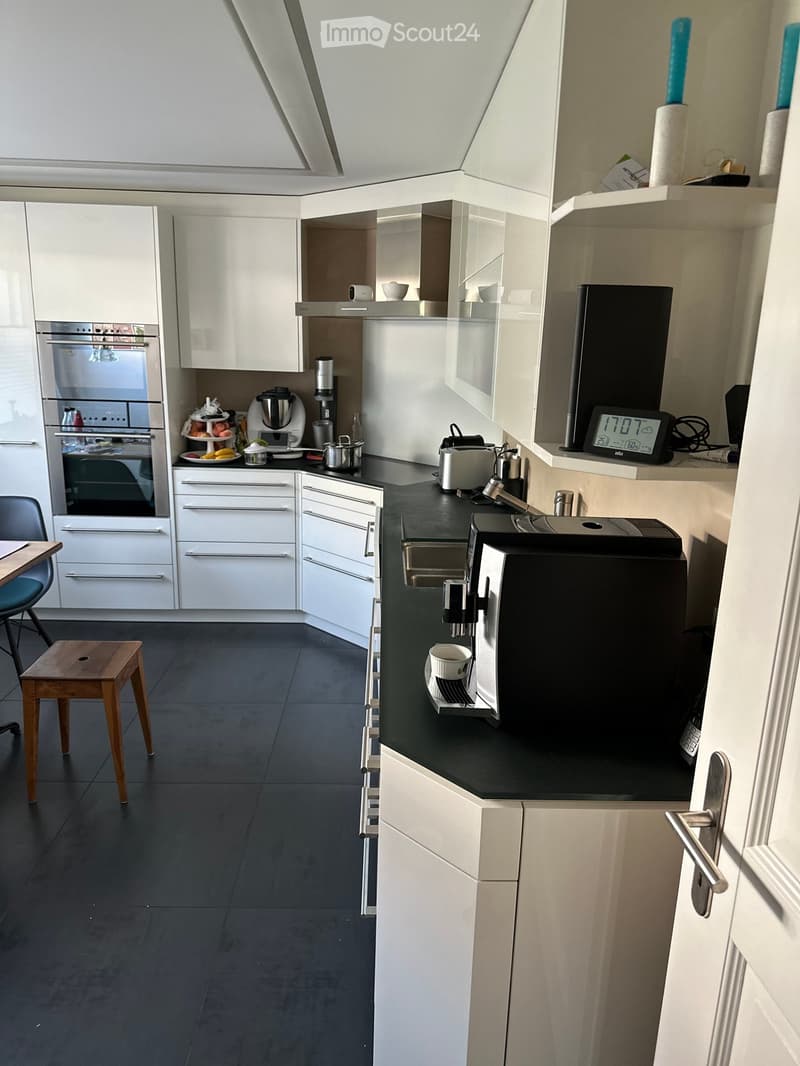 Modern kitchen with white cabinets, black countertops, and stainless steel appliances. Includes a coffee machine, blender, oven, sink, and a small wooden stool.