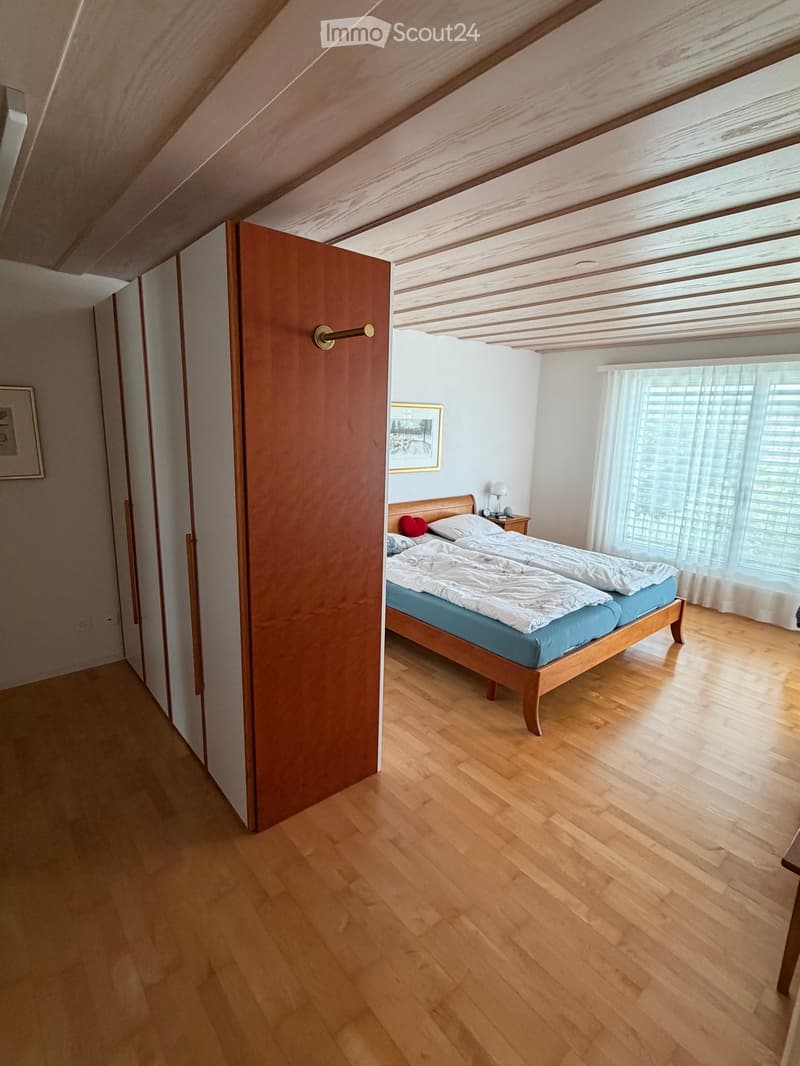 Bright bedroom with wooden floors, a double bed with white bedding, a nightstand with a lamp, and a picture frame on the wall. The room features large windows with blinds and a slanted wooden ceiling.