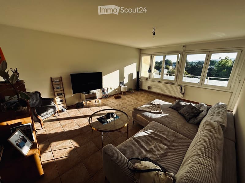 Bright living room with large windows, tiled floor, sofa, coffee table, armchair, TV, and decorative items.