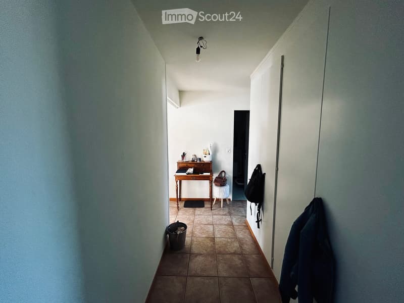 A narrow hallway with tiled flooring, a small wooden table with a stool, a handbag on the stool, a backpack hanging on the wall, and a coat draped over the wall.
