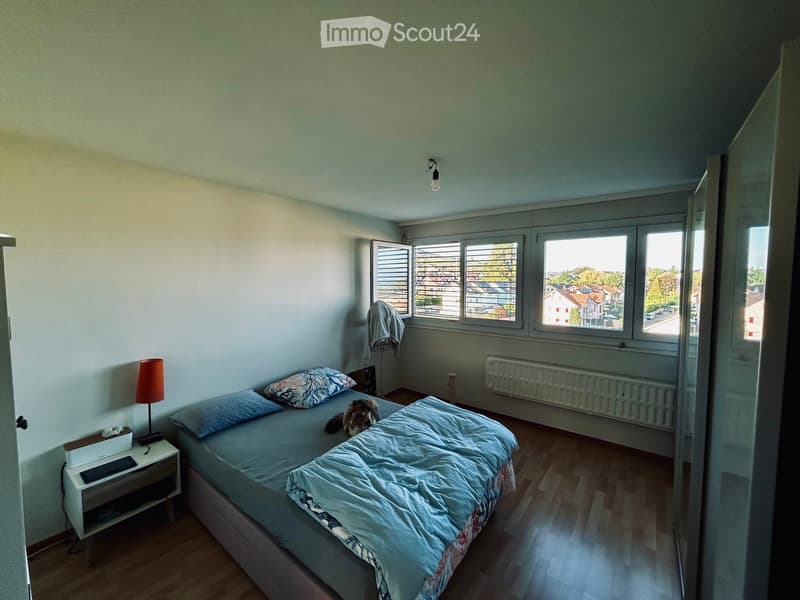 Bedroom with a double bed, nightstand, lamp, radiator, and a view of the neighborhood through large windows.
