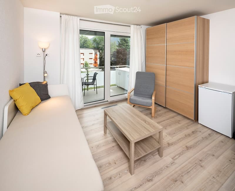 Cozy studio with a sofa, small table, chair, wardrobe, and refrigerator. Large window with balcony access.