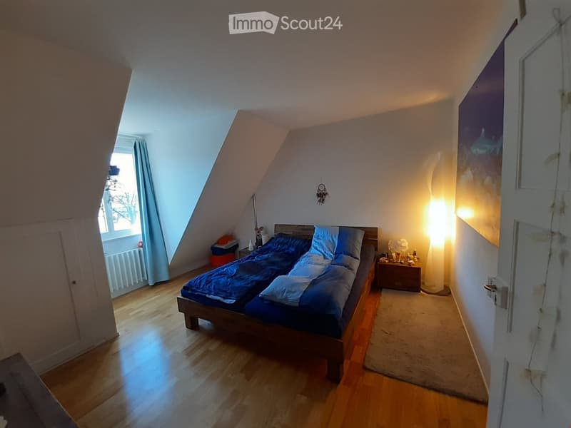 Cozy attic bedroom with wooden floors, slanted ceilings, large window with blue curtains, double bed with blue bedding, nightstand, floor lamp, and decorative wall art.