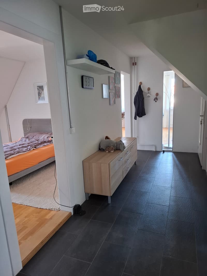 Modern bedroom with a slanted ceiling, dark tiled floor, and wooden flooring near the bed. Features a bed with orange bedding, a nightstand, a wooden drawer unit with toys, framed pictures on the wall, a clock, and a coat rack with a jacket.