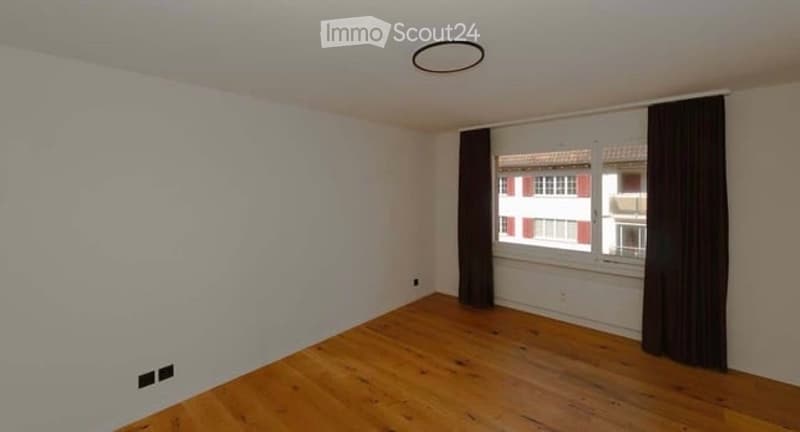 Empty room with wooden flooring, white walls, and a window with dark curtains. No furniture or equipment present.