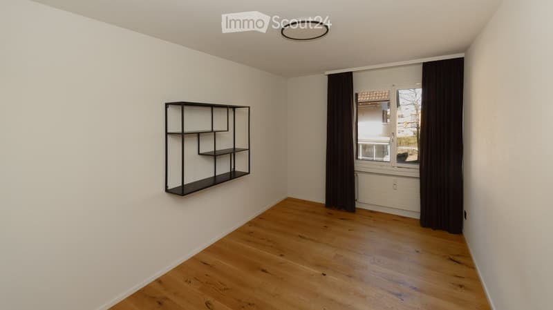 Empty room with wooden flooring, white walls, a black shelving unit, and a window with dark curtains.