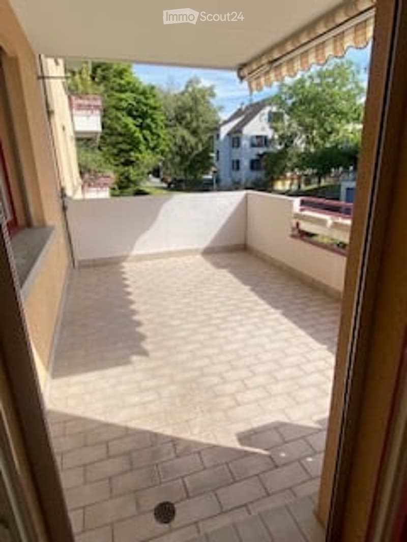 Balcony with brick floor, white railing, and striped awning. Offers view of greenery and neighboring buildings.