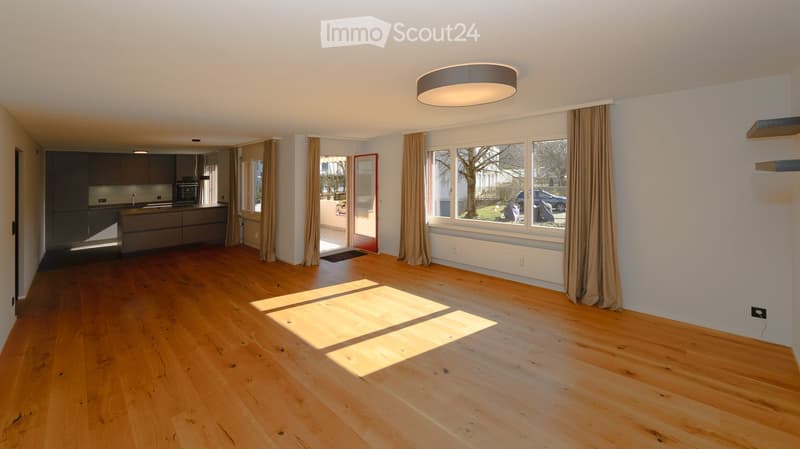 Open-plan living and kitchen area with wooden flooring, modern kitchen cabinets, and large windows with curtains.