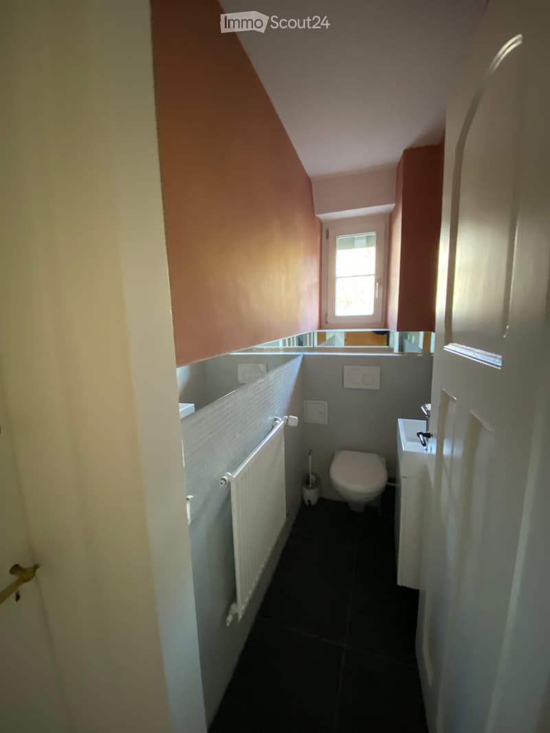 Small bathroom with a toilet, sink, radiator, and toilet brush. The walls are tiled, and there is a window at the far end.