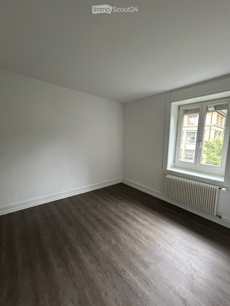Empty room with light grey walls, dark wooden flooring, and a single window with white frame and radiator below it.
