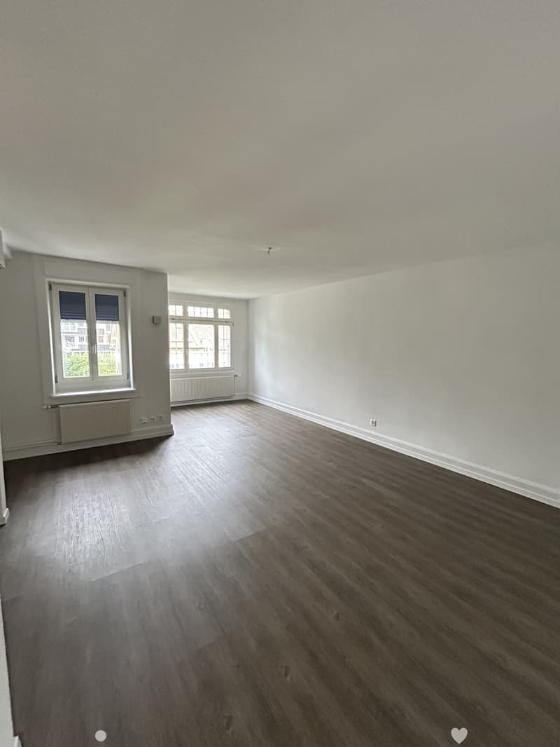 Empty room with wooden flooring, white walls, and large windows with radiators below them.