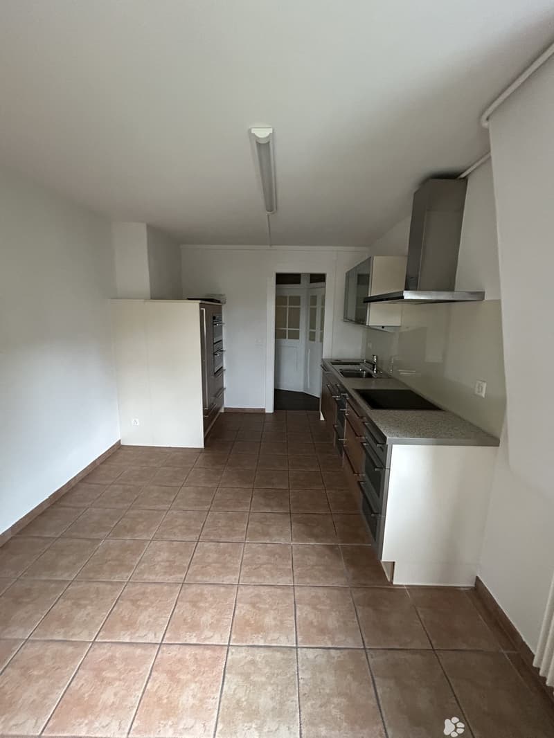 Modern kitchen with tiled floor, white cabinets, stainless steel sink, and oven. Features a built-in range hood and a doorway leading to another room.