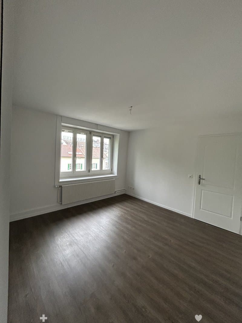 Empty room with light-colored walls, wooden flooring, a single window with white frames, and a white door.
