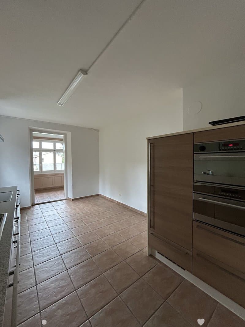 Modern kitchen with tiled floor, built-in oven, and wooden cabinets. A doorway leads to a balcony area with wooden furniture.
