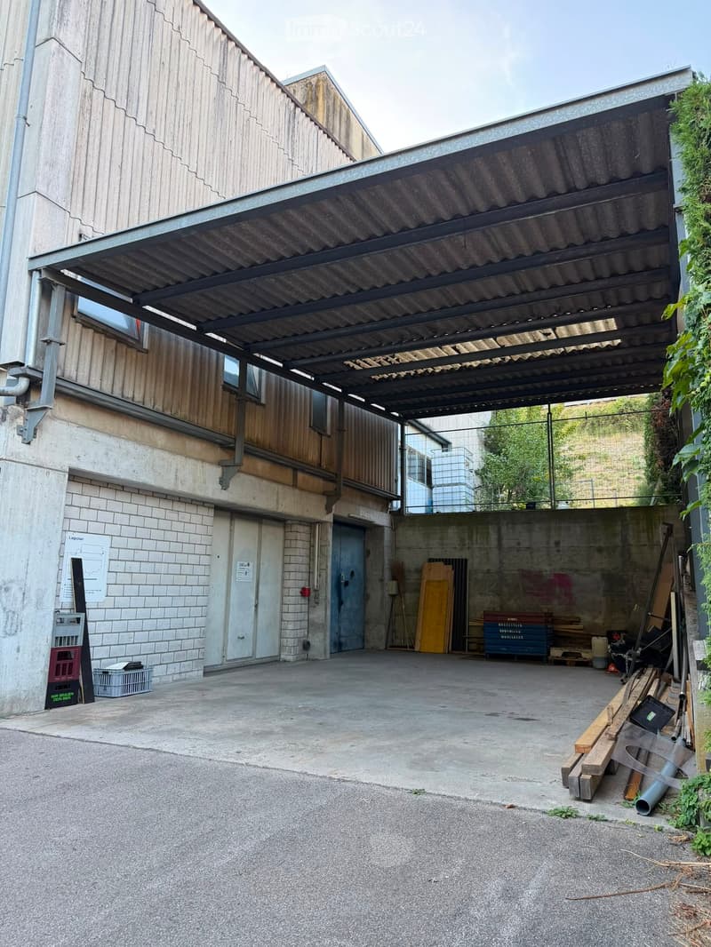 A covered outdoor area with a metal roof structure, concrete floor, and various stored items including wooden planks, crates, and tools.