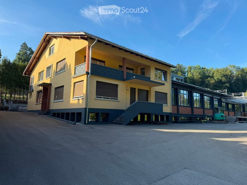 Two-story yellow building with a raised ground floor, multiple windows, and a balcony on the upper level.