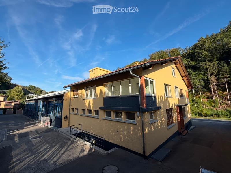Two-story yellow house with a red-tiled roof, multiple windows, and a small balcony on the upper floor. The house is situated on a raised ground floor with a staircase leading up to the entrance.