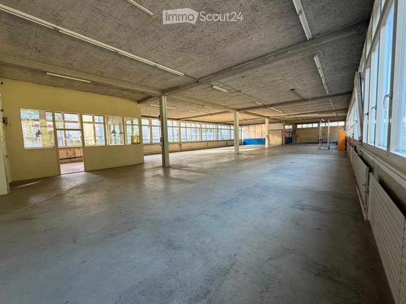 Large, empty industrial-style room with concrete floors, exposed ceiling beams, and large windows allowing natural light. Minimal furnishings, with a few scattered items like a ladder and small cabinets visible in the background.