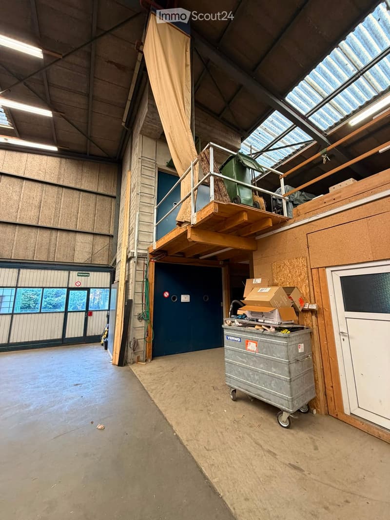 Empty industrial-style room with concrete floor, exposed wooden beams, and large windows. Features a metal staircase leading to an upper level, a large blue door, and a grey wheeled container with boxes on top.