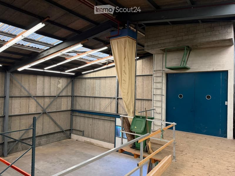 Empty industrial-style room with exposed metal beams, concrete walls, and a large blue door. Features include a metal staircase, a green waste bin, and a partially covered column.