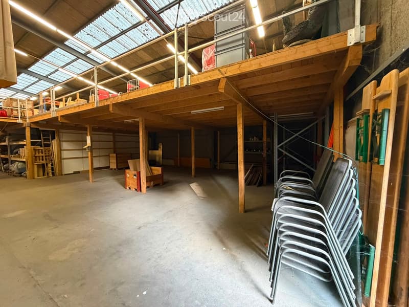 Large, open industrial-style space with wooden beams, concrete floor, and a raised platform with storage. Stacks of metal chairs are positioned against the wall.