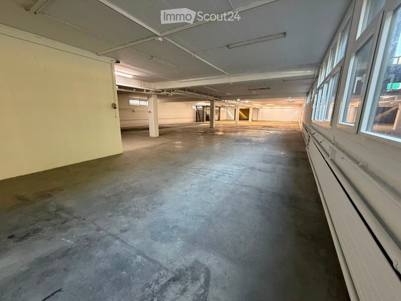 Empty corridor with concrete floor, white walls, and large windows on one side. Exposed ceiling with fluorescent lighting and structural beams.
