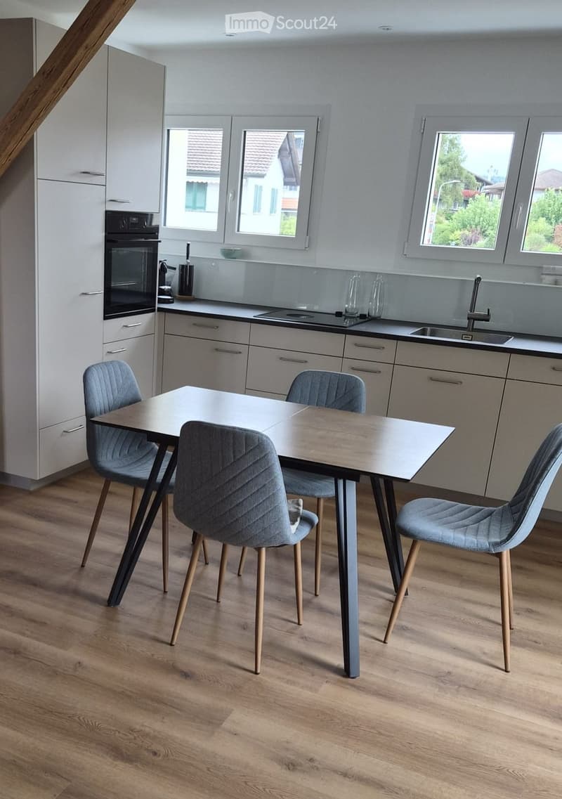 Modern kitchen with light grey cabinets, a central dining table with four grey upholstered chairs, and a built-in oven. The kitchen features a sink with a modern faucet and ample natural light from large windows.