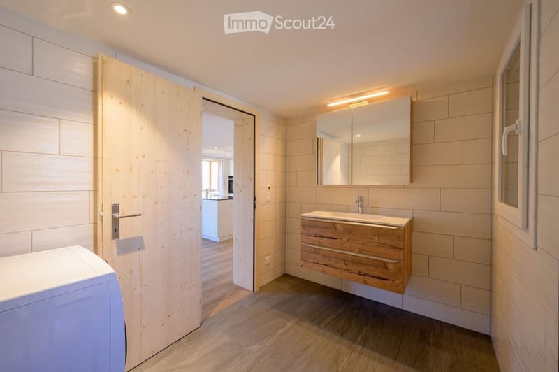 Modern bathroom with light wooden cabinet, sink, and mirror. The room features light-colored tiles and a wooden door leading to another space.