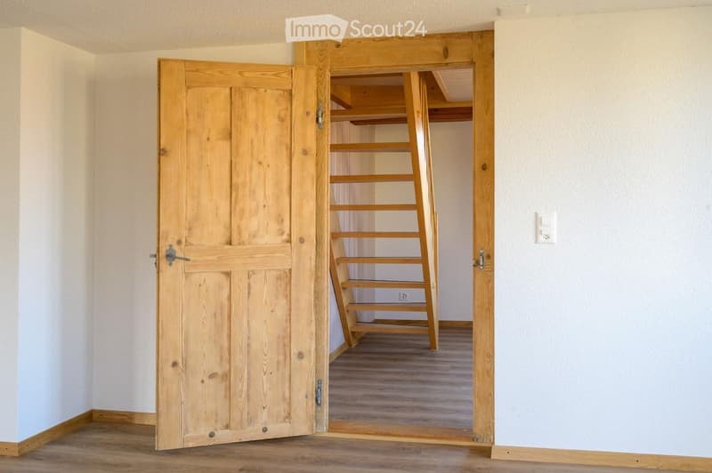An empty room with wooden flooring and white walls, featuring an open wooden door leading to a closet with wooden shelves and a small ladder.