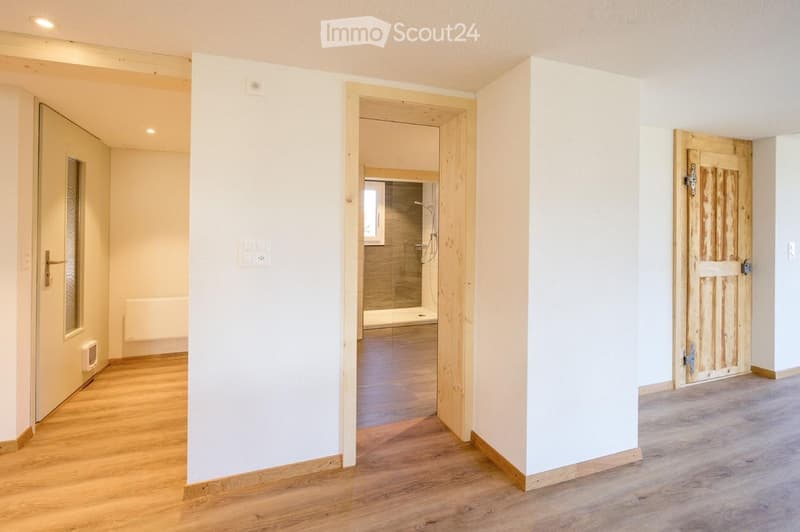 Empty hallway with wooden flooring, white walls, and multiple doors leading to different rooms, including a bathroom with a glass shower enclosure.