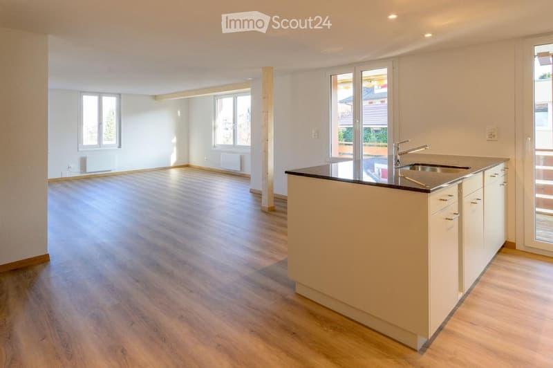 Empty kitchen with light wooden flooring, white walls, and a modern kitchen island with a sink and faucet. Large windows provide natural light.