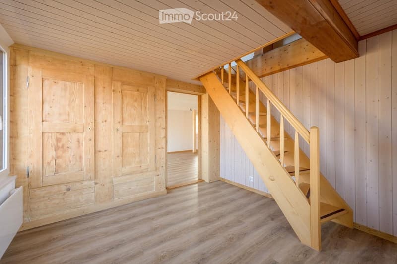 Empty room with wooden walls, wooden floor, and a staircase leading upwards. The space is clean and unfurnished, with natural light coming through a window on the left side.