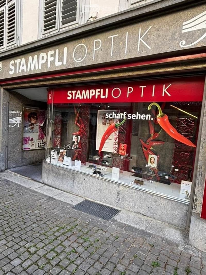 Storefront of an optician shop named "Stampfli Optik" with a red awning, glass display case, and various optical products visible inside.