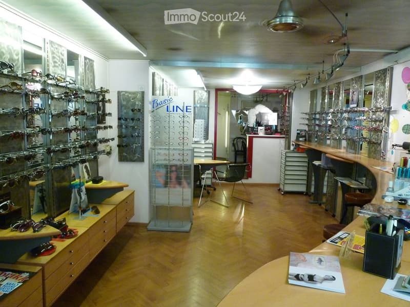 A well-lit optical shop with wooden flooring, display racks filled with sunglasses, a counter with brochures, and seating areas for customers.