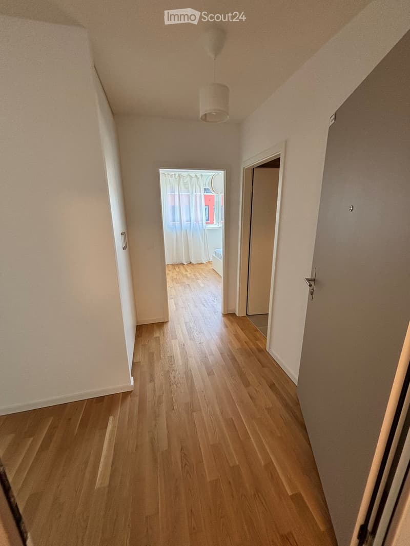 A narrow hallway with wooden flooring, white walls, and a hanging light fixture. There are two doors on either side, one leading to a room with a window and curtains.