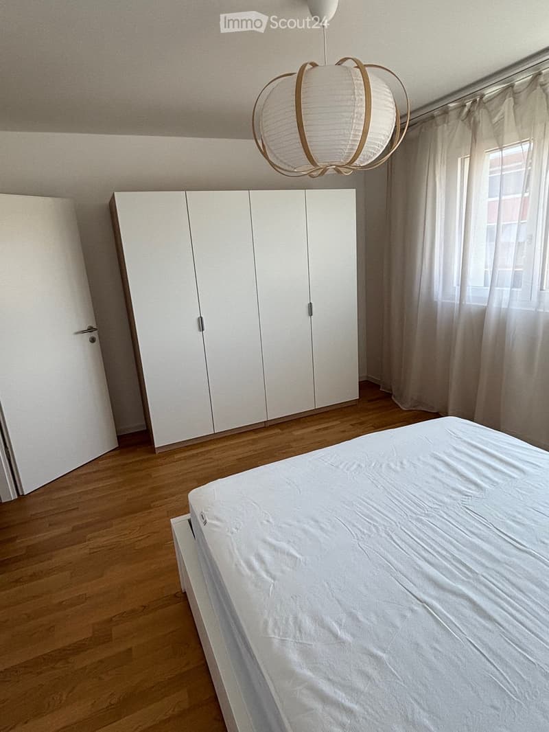 A modern bedroom with a white bed, large white wardrobe, wooden flooring, and a hanging light fixture.