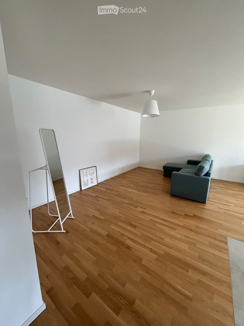 A modern, minimally furnished room with wooden flooring, white walls, a grey sofa, a mirror on a stand, and a decorative picture frame that reads "HOME".