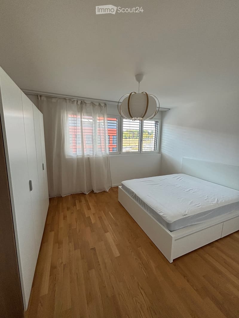 Minimalist bedroom with a white bed, wooden floor, large window with curtains, and a modern hanging light fixture.