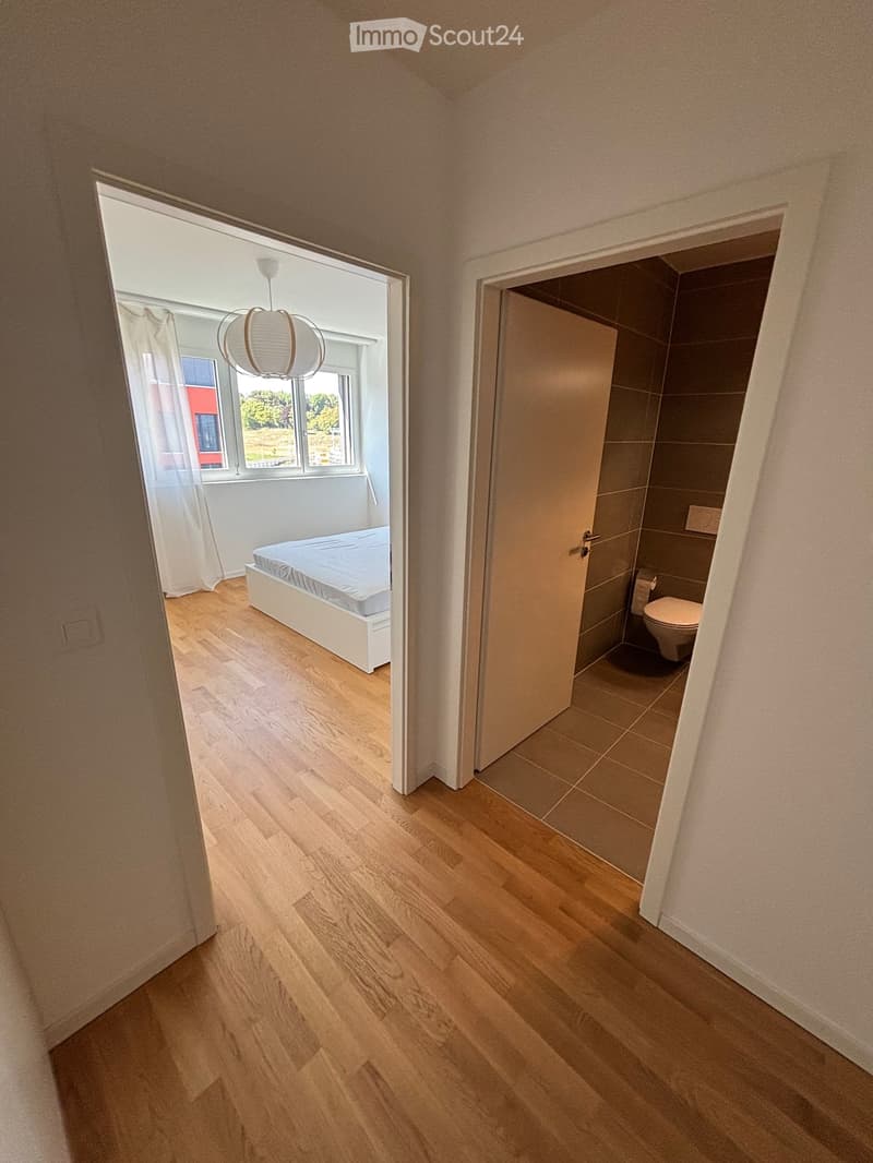 A small, modern bedroom with light wooden flooring, a white bed frame, and a partially open door leading to a bathroom with a toilet.