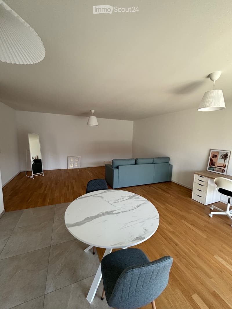Modern living room with wooden flooring, a grey sofa, a white marble table, and a grey chair. There is a mirror, a lamp, a picture frame, and a desk with a chair.