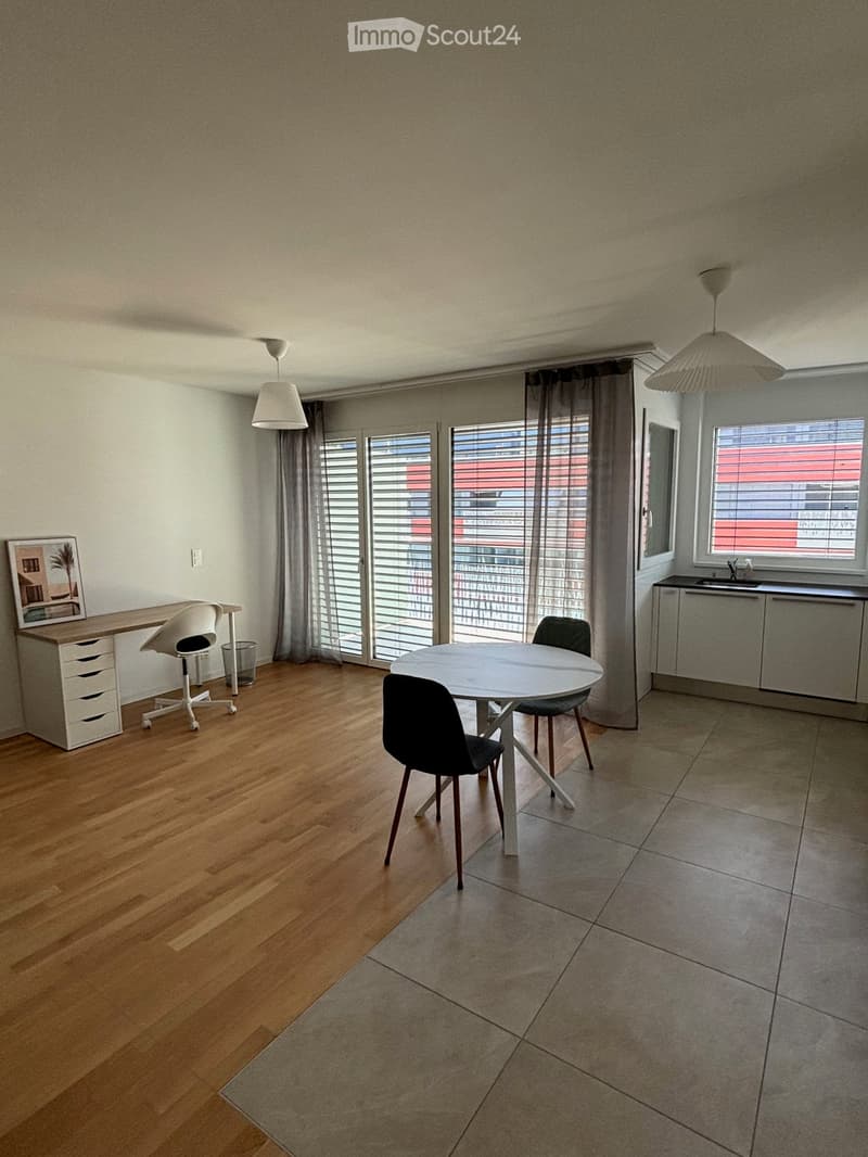 A modern, open-plan room with a dining area and a workspace. Features include a round dining table with chairs, a desk with a chair, a small kitchen area with a sink and cabinet, large windows with blinds, and pendant lights.