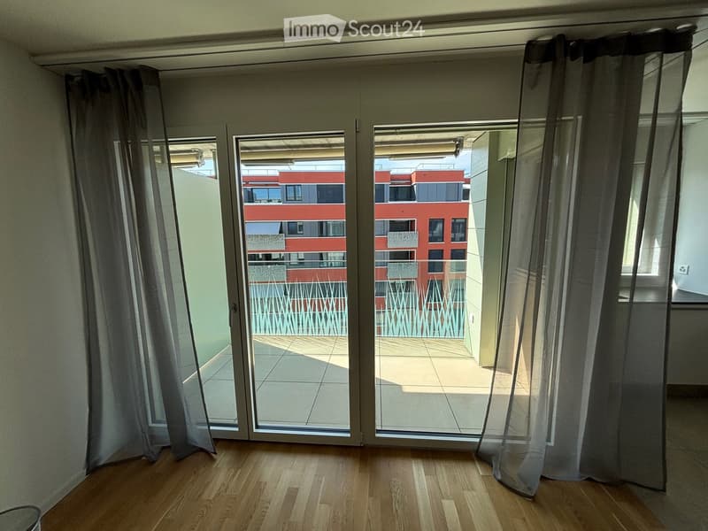 Empty room with wooden flooring, large sliding glass doors leading to a balcony, and grey curtains on either side.