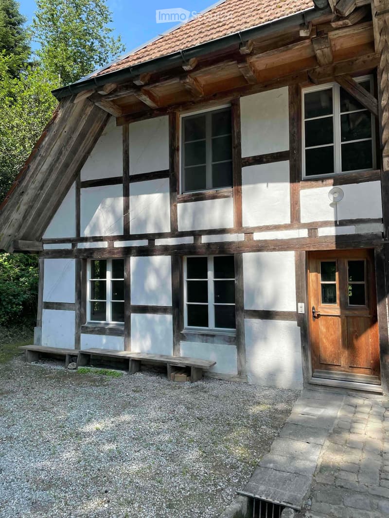 Traditional half-timbered house with white plaster walls, wooden beams, multiple windows, and a wooden front door. Gravel driveway and wooden benches are visible in front.