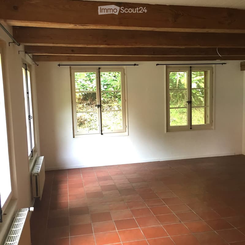 Empty room with exposed wooden beams on the ceiling, red tiled floor, and multiple windows with shutters. Bare walls and no furniture.