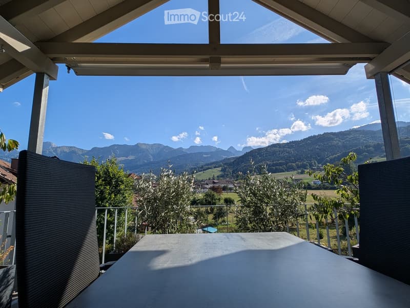A covered balcony with a large table, overlooking a scenic mountain landscape with trees and a village below.