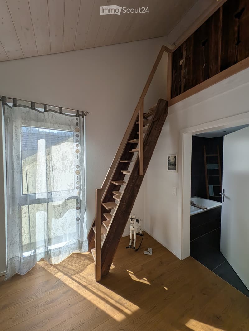 Slanted wooden staircase leading up, with a small toy horse and heart-shaped decoration on the floor, a window with sheer curtains, and a view into a bathroom with a sink and bathtub.